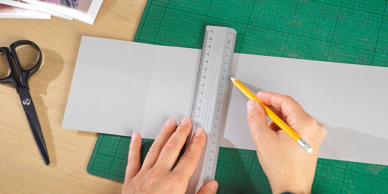 Dvije ruke olovkom i ravnalom crtaju linije za fotografije na listu sivog kartona.